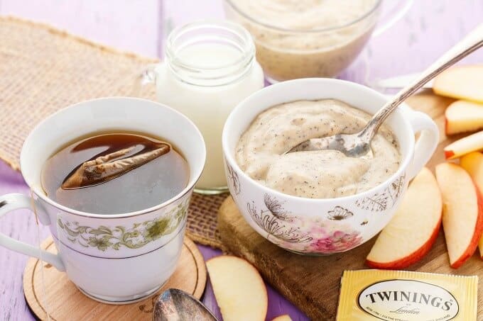 Homemade Earl Grey Pudding in cups with spoon, wooden pad with apple slices, cup of tea, jar of milk on purple table