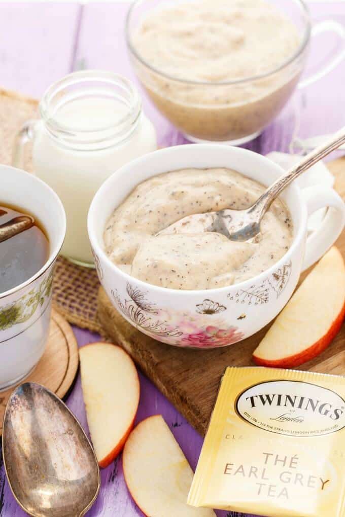 Homemade Earl Grey Pudding in white cups with spoon on wooden pad with slices of apple, jar of milk, spoon, tea bag, cup of tea on purple table