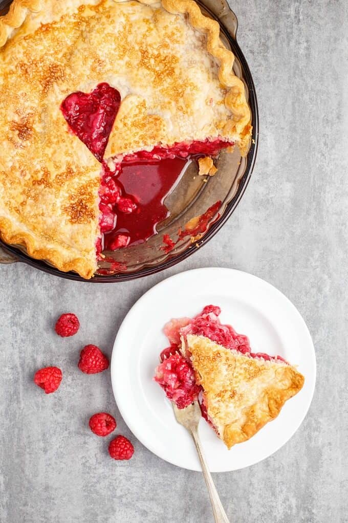 Homemade Raspberry Pie in baking pot, slice of pie on white plate with fork. Scattered raspberries on gray table