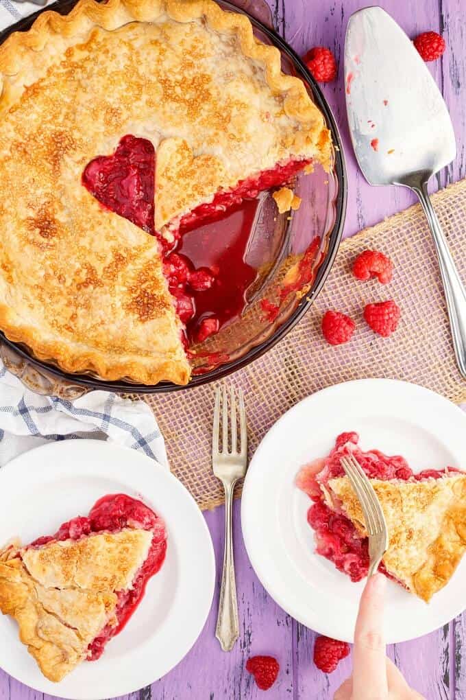 Homemade Raspberry Pie in baking pot and slices of pie on white plates. Fork held by hand, fork, raspberries scattered on table, spatula and cloth wipe on purple table.