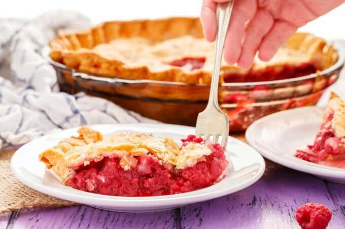 Homemade Raspberry Pie on white plates with fork held by hand. Cloth wipe, rest of the  cake in baking pot and and raspberrey on purple table