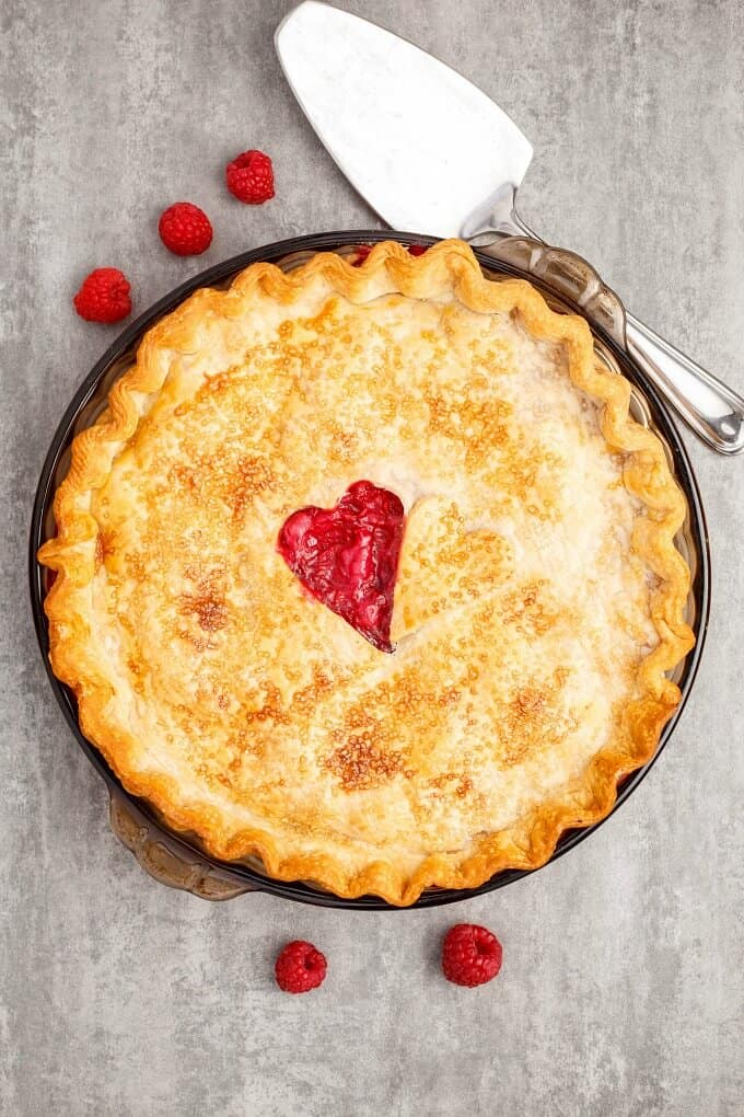 Homemade Raspberry Pie in baking pot with scattered raspberries and spatula on gray table.