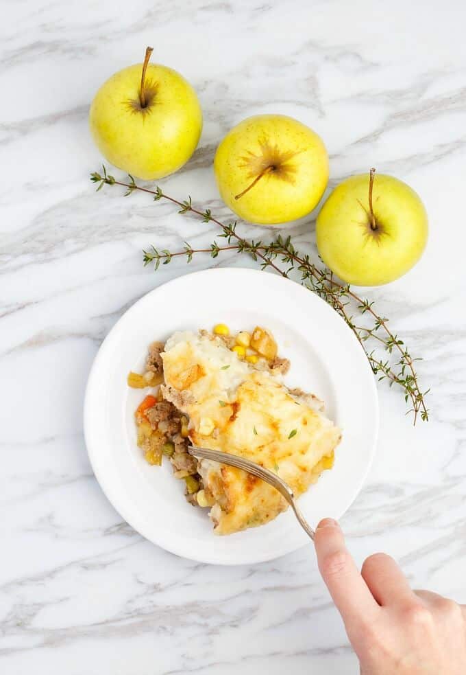 Apple-Pork Shepherd's Pie in white plate with fork held by hand. Herbs, apples on white-gray table