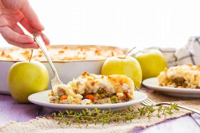 Apple-Pork Shepherd's Pie in baking pot, on white plates with fork held by hand. Fork, herbs, green apples and cloth wipe on purple table