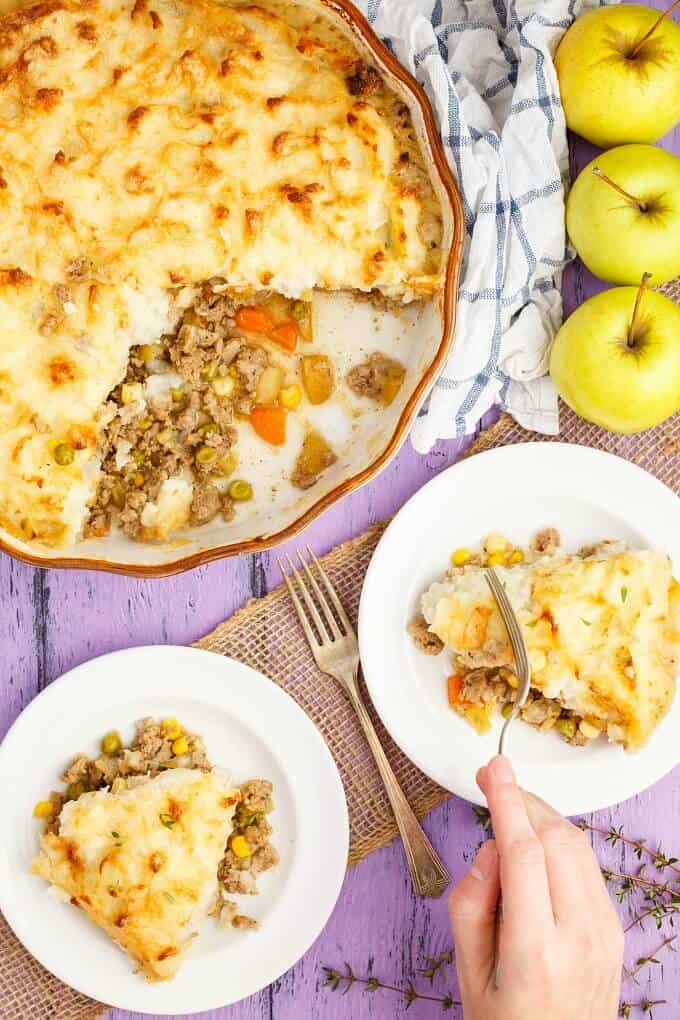 Apple-Pork Shepherd's Pie in baking pot and on white plates with fork held by hand. Fork, apples, herbs and cloth wipe on purple table