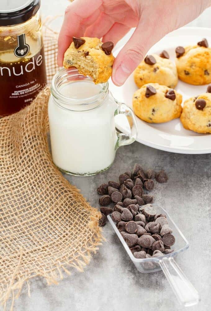 Honey Chocolate Chip Cookies  on white plate and over jar of milk held by hand. Brown cloth, jar of honey and spatula with chocolate chips on gray table