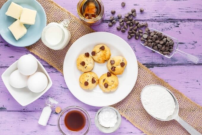 Honey Chocolate Chip Cookies  on white plate with jars of milk and honey, Bowls of ingredients, bowls of eggs and butter. spatulss with flour and chocolate chips on purple table