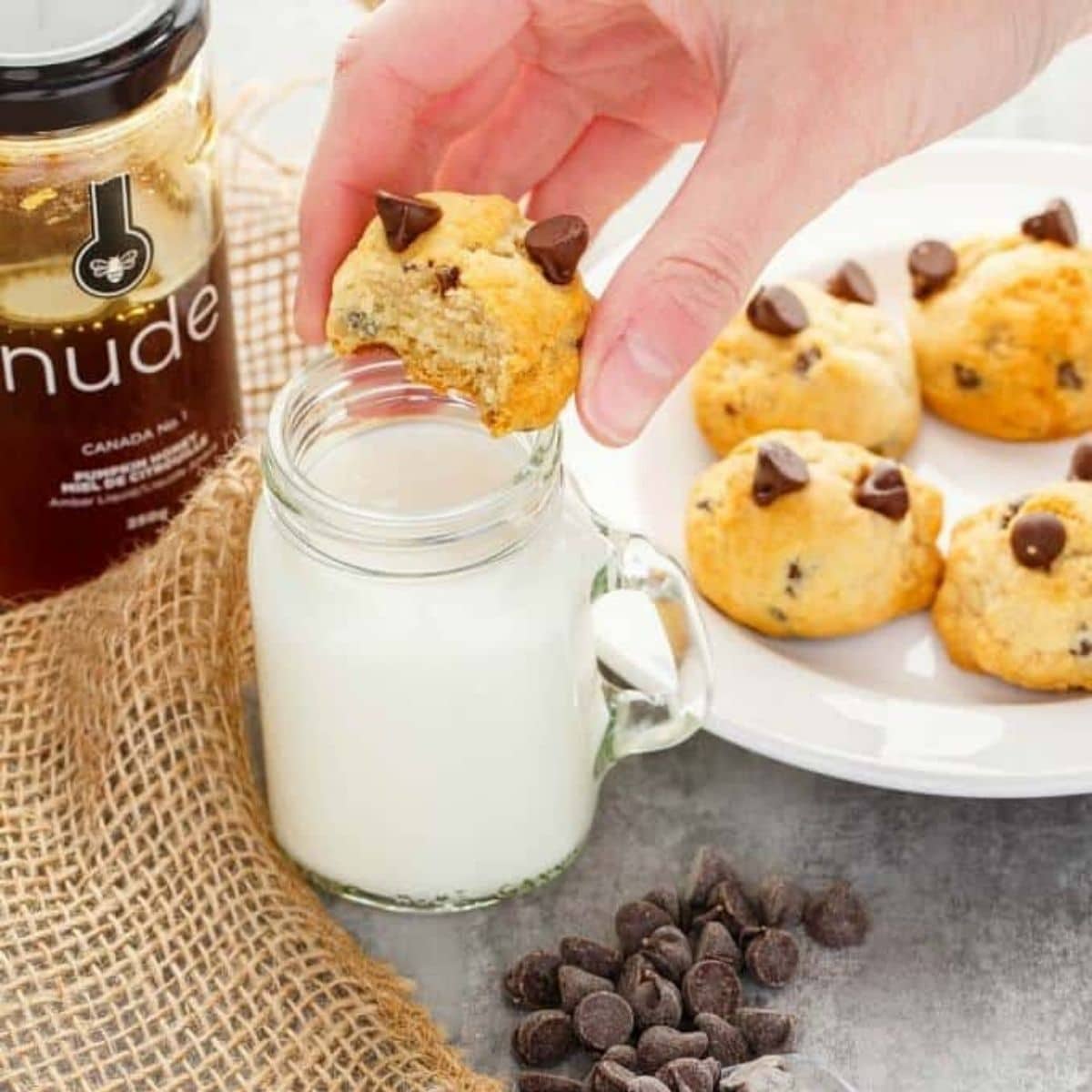 Honey chocolate cip cookes on white platem one cookie held by hand over glass of milk, with chocolate chips and jar of honey on table