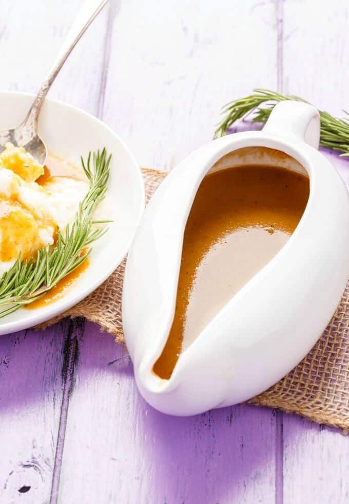 Homemade Turkey Gravy in white bowl next to white plate with dish, herbs and spoon. Herb, brown cloth on purple table