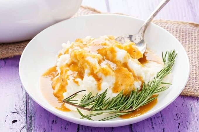 Homemade Turkey Gravy on white plate with mashed dish, herb and spoon. Brown cloth, white bowl on purple table