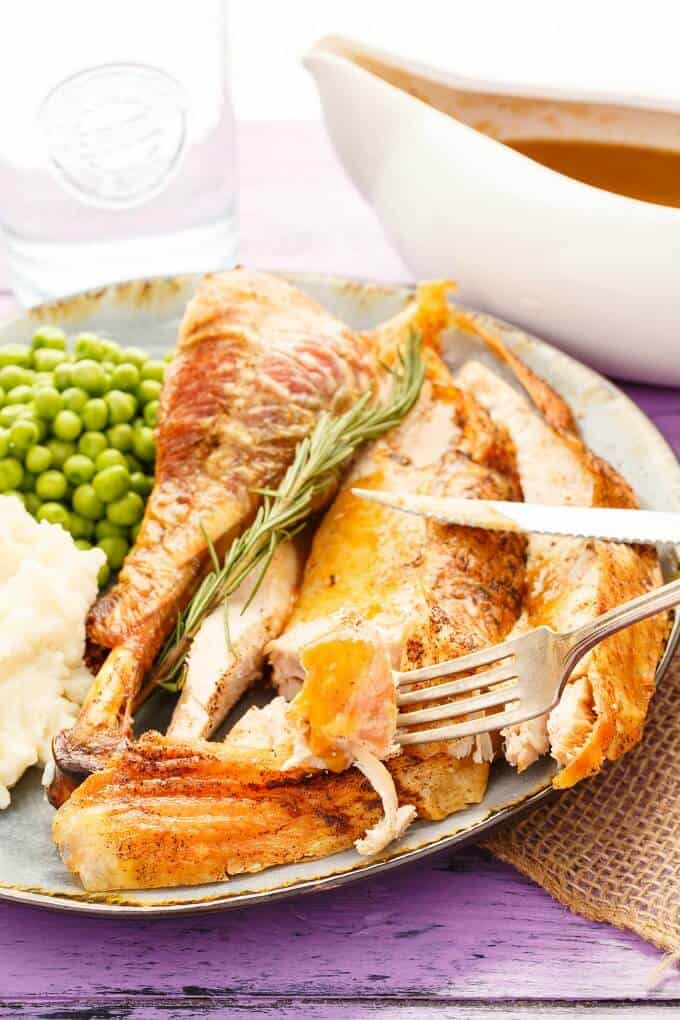 Oven Spatchcock Turkey  on blue plate with knife, fork, mashed potatoes and peas. Blue bowl with sauce, glass cup, brown cloth on purple table