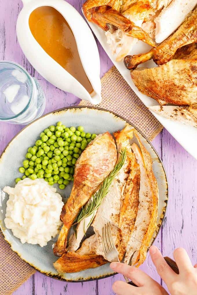 Oven Spatchcock Turkey  on blue plate with peas, mashed potatoes, herb, knife and fork held by hand. Turkey on white tray, White bowl of sauce, glass cup, brown cloth on purple table