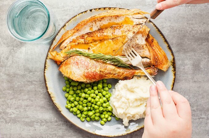 Oven Spatchcock Turkey  on blue plate with peas, mashed potatoes, herbs, fork and knife held by hand. Glass cup on gray table