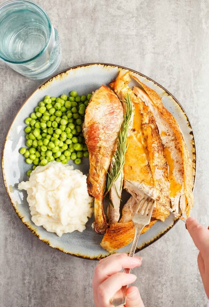 Oven Spatchcock Turkey on blue plate with herb, peas, mashed potatoes, fork and knife held by hands with glass of water on gray table