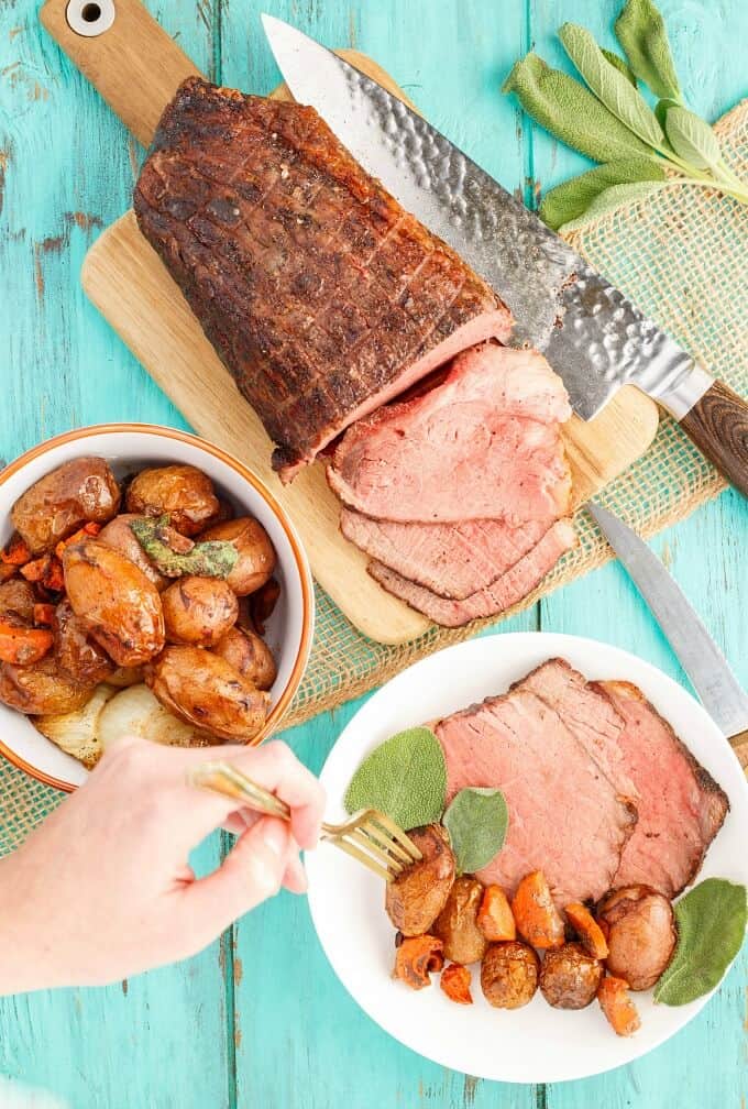 Sage Butter Round Roast with Creamer Potatoes on white palte with fork being held by hand. Bowl of potatoes, roung on wooden pad partialy sliced, knifes, herbs and brown cloth on blue table