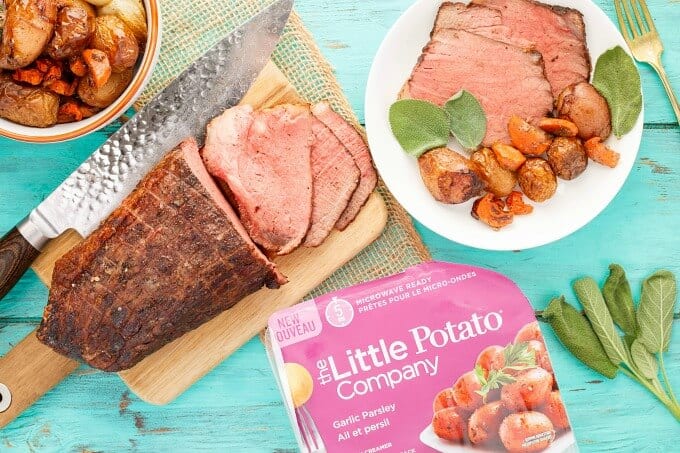 Sage Butter Round Roast with Creamer Potatoes on white plate. Wooden pad with roasted round beef, knife, fork, herbs, bowl of potatoes and potato package on blue table