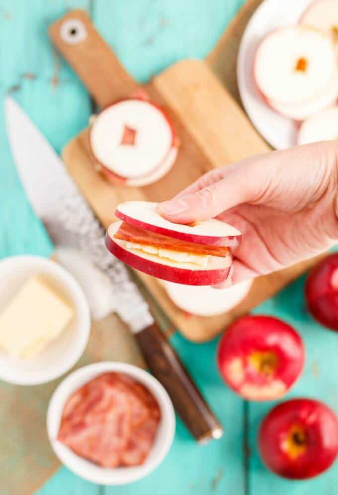 Turkey Bacon Apple Sandwich with Cheese held by hand over wooden pad, knife, apples, bowls of ingredients on blue table