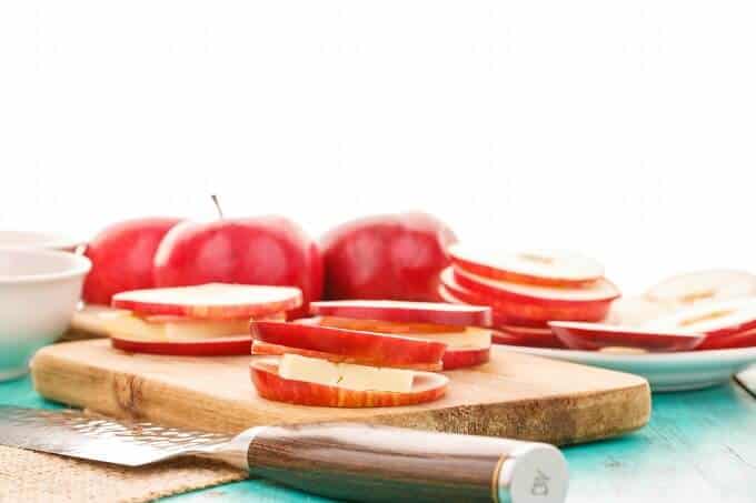 Turkey Bacon Apple Sandwiches with Cheese on wooden pad. Knife, red apples, white bowl, white plate with slices of apple on blue table