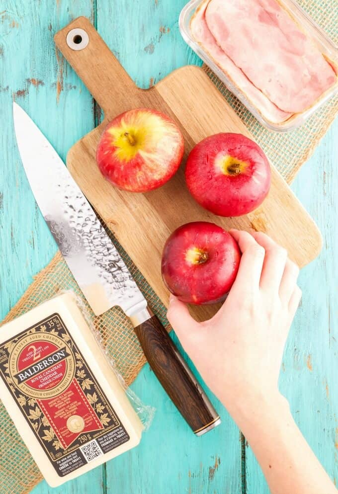 Red apples touched by hand on wooden pad with knife, pork bacon slices, cheese in container, brown cloth on blue table