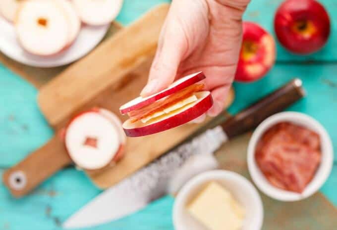 Turkey Bacon Apple Sandwich with Cheese held by hand over wooden pad witch sandwich, knife, bowl of bacon and cheese, red apples and sliced apples on white plate on blue table