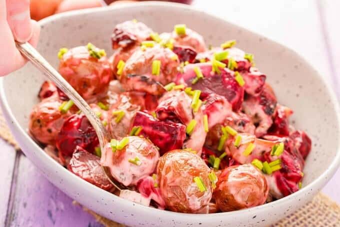 Roasted Beet Potato Salad in whitish bowl with spoon held by hand on purple table