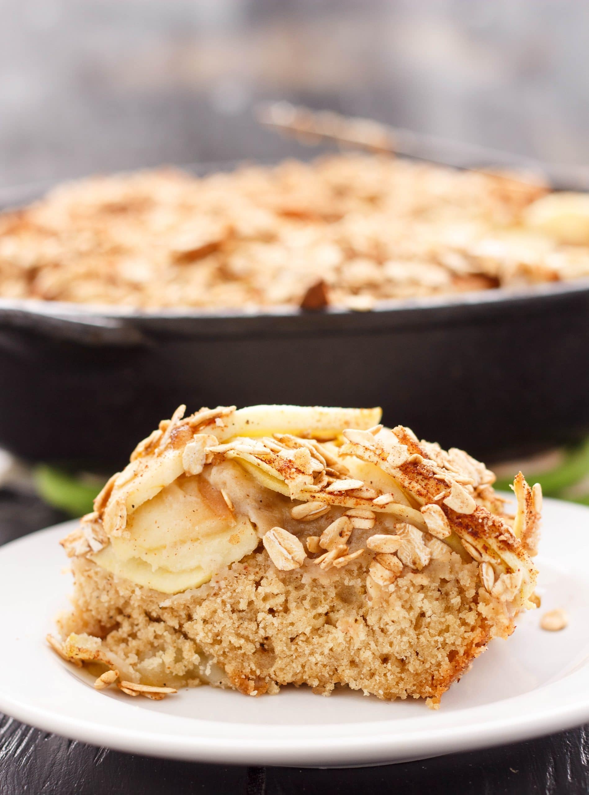 Skillet Apple Cake with Cinnamon Oat Topping on white plate, black pan with cake in the background