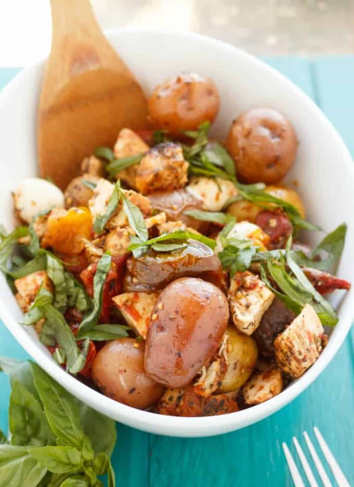 Flavorful Roasted Potato and Chicken Caprese Salad in a white bowl with a wooden spatula.